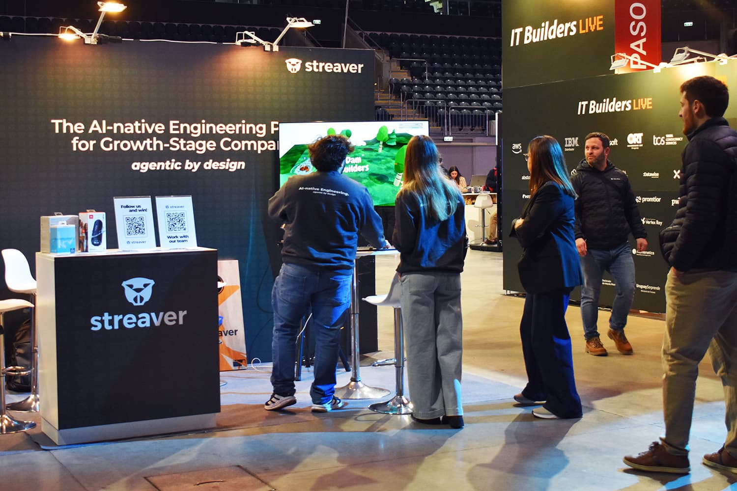 Two visitors play Dam Builders on the stand's screen, next to the prizes and the Follow us and win QR codes