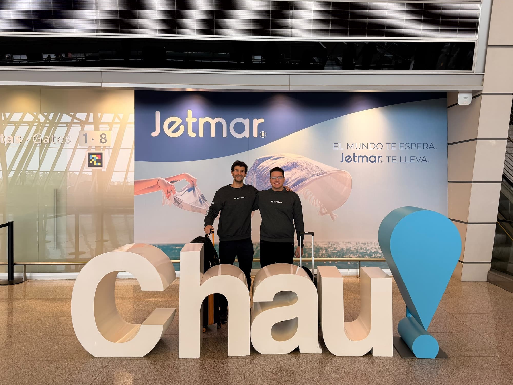 Fabián Larrañaga and Agustín Tornielli in Streaver sweatshirts, with their suitcases, behind a giant "Chau!" sign at the airport