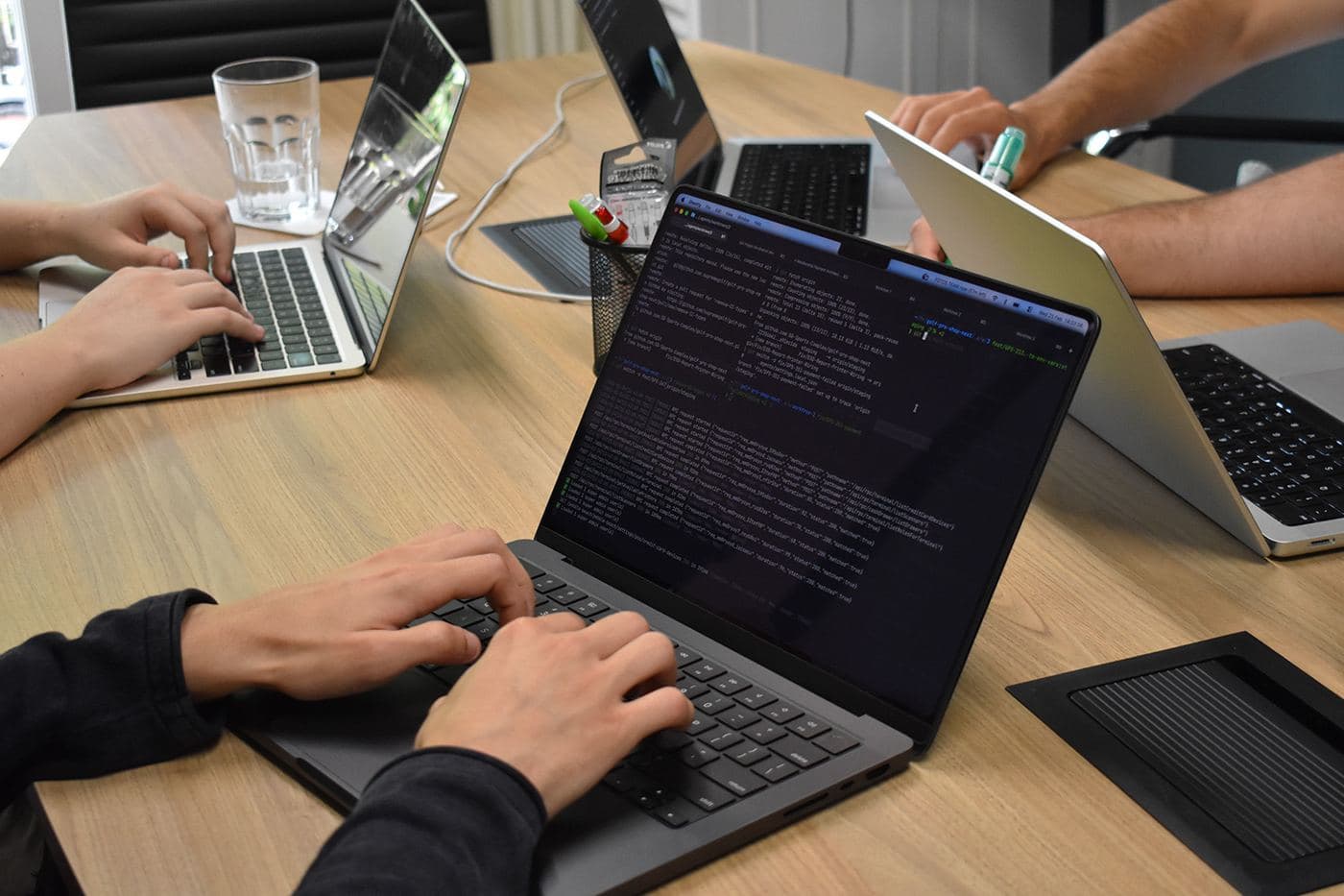 Hands on laptops around the office table, a terminal full of code on the nearest screen