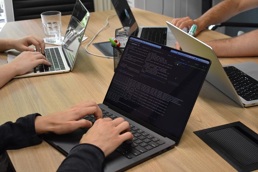Hands on laptops around the office table, a terminal full of code on the nearest screen