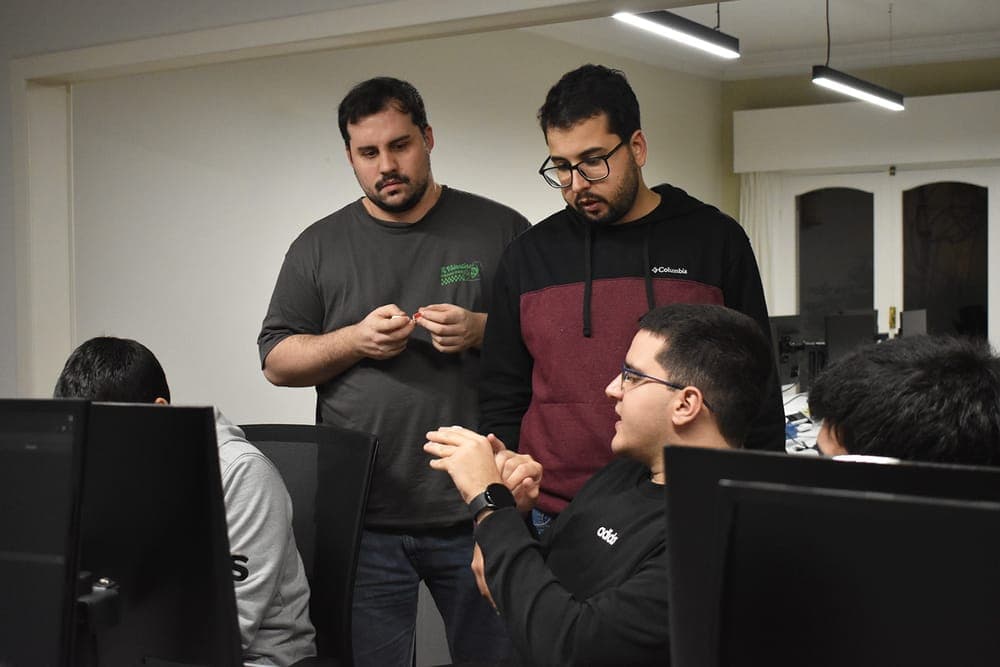 Two teammates standing over a colleague's desk, talking through what is on his screen at the hackathon